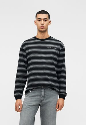 Jonge man die een zwart-grijs gestreept shirt met lange mouwen en tekst op de borst draagt, met lichtgrijze jeans, staand tegen een effen achtergrond.