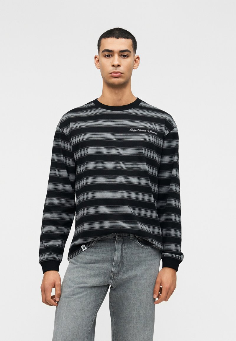 Νέος άνδρας που φοράει μαύρο και γκρι ριγέ μακρυμάνικο πουκάμισο με κείμενο στο στήθος και ανοιχτό γκρι τζιν, στέκεται μπροστά σε απλό φόντο.