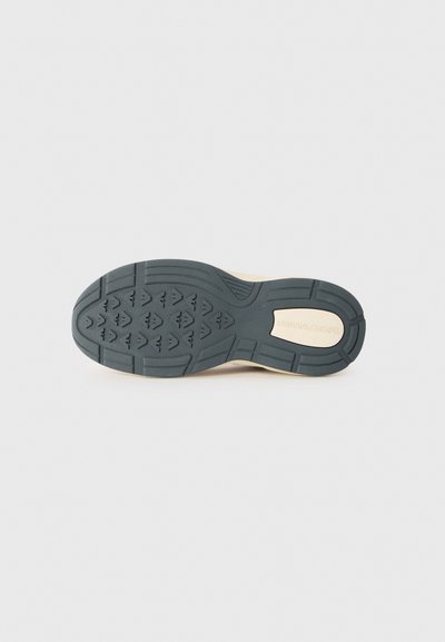 Sålen av en beige sneakersko med svart mønstret gummisåle og synlig merkevare navn preget på den beige hældelen mot hvit bakgrunn.