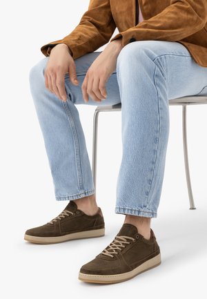 Man die lichtblauwe spijkerbroek, bruine suède sneakers met beige zolen en een bruine jas draagt, zittend op een metalen stoel.