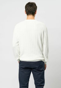 Bărbat purtând un pulover alb texturat cu model împletit și pantaloni bleumarin închis, stând cu spatele spre un fundal simplu, gri deschis.