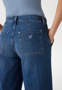 Dunkelblaue Denim-Jeans mit taillierter Passform, zwei Gesäßtaschen mit blauem Dreieckslogo und einer glatten Textur.