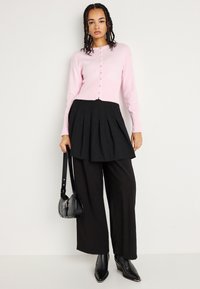 Ανοιχτό ροζ πλεκτό ζακέτα με κουμπιά, μαύρη πλισέ φούστα, φαρδιά μαύρα παντελόνια και μυτερές μαύρες μπότες αστραγάλου; περιλαμβάνει μια μαύρη τσάντα ώμου.