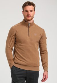 Tan zip-up fleece pullover met een hoge kraag, textuurstof en een zijzak. Voorzien van een logopatch op de borst en geribde manchetten.
