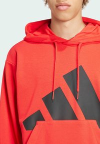 Felpa rossa in cotone con tasca frontale, caratterizzata da un grande design di logo nero. Include lacci regolabili sul cappuccio e polsini a coste.