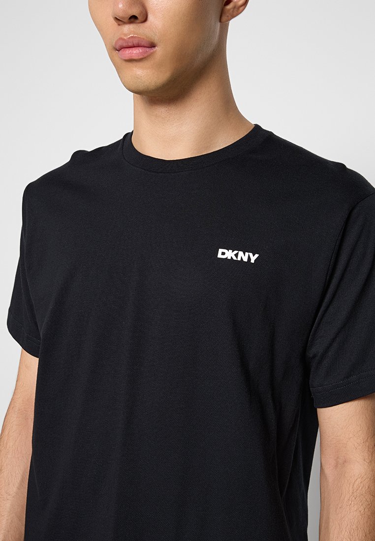 Camiseta de algodón negra con cuello redondo y mangas cortas, que presenta un logo blanco de DKNY en el lado izquierdo del pecho. Textura suave, ajuste clásico.