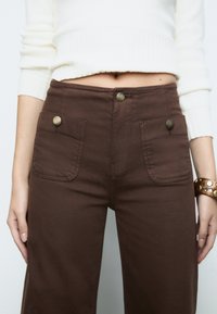 Brown cotton pants with two front pockets, metal buttons, and a slightly textured fabric; worn with a cropped white sweater.