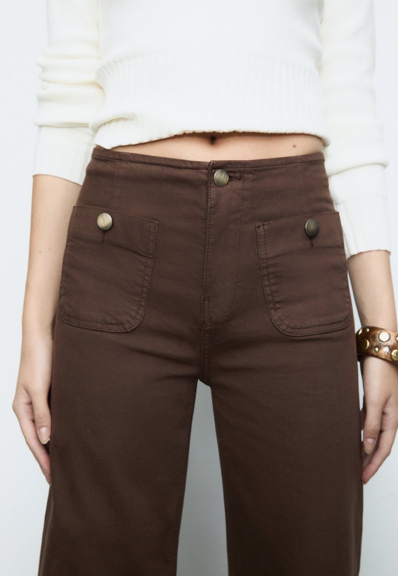 Brown cotton pants with two front pockets, metal buttons, and a slightly textured fabric; worn with a cropped white sweater.