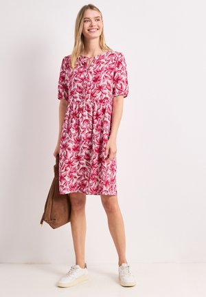 Femme souriante portant une robe rose fleurie mi-longue, des baskets blanches, et tenant un sac en daim marron sur un fond clair uni.