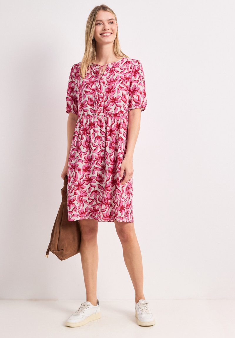 Femme souriante portant une robe rose fleurie mi-longue, des baskets blanches, et tenant un sac en daim marron sur un fond clair uni.