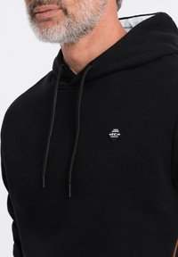 Sudadera negra con capucha que presenta un forro gris, cierres de cordón y un pequeño parche negro con logo en el pecho. Textura de tela suave.