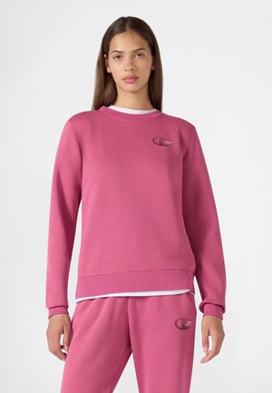 Joven mujer vestida con sudadera y pantalones deportivos rosas a juego, de pie frente a un fondo blanco liso.