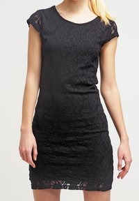 Kvinna klädd i en figurnära, kort svart spetsklänning med korta ärmar, stående mot en enkel ljusbakgrund.