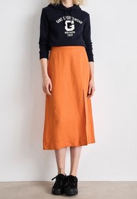 Personne portant un sweat à capuche noir avec un texte blanc et une jupe midi orange fendue à l'avant, associée à des baskets noires, debout contre un mur blanc.