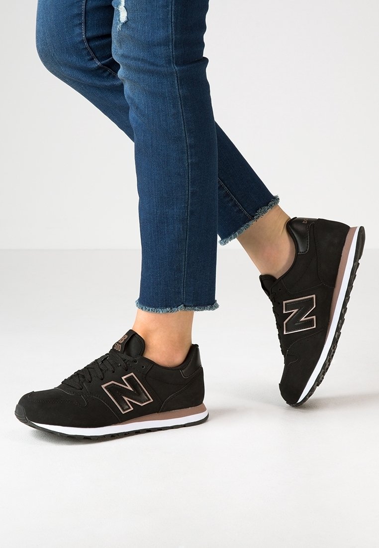 Personne portant des baskets New Balance noires et un jean skinny bleu avec ourlet effiloché, debout sur un sol blanc.
