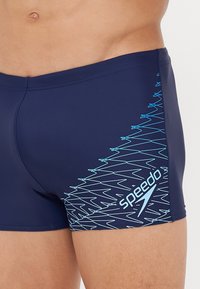Tengerészkék fürdőnadrág, rugalmas anyagból készült, kék cikk-cakk mintával és Speedo logóval a bal oldalon.