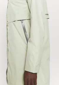 Ljusgrön vattentät jacka med en slät yta, utrustad med en ficka på sidan med dragkedja och en logotyp på ärmen. Modern design med minimala detaljer.