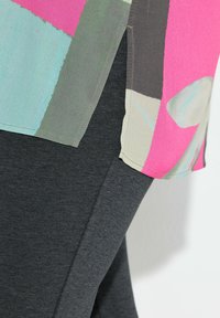 Farverig bluse med rosa, grønne og grå mønstre, lagdelt over mørkegrå leggings. Blusen har sidesplitter og en blød tekstur.