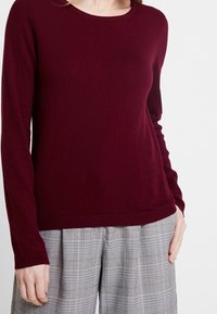 Bordeaux lange mouwen trui met ronde hals, zachte textuur en geribbelde manchetten. Gecombineerd met grijs geruite hoge taille broek.