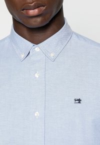 Lichtblauw overhemd van katoen met een klassieke kraag, witte knopen en een klein zwart naaimachine-logo op de borst.