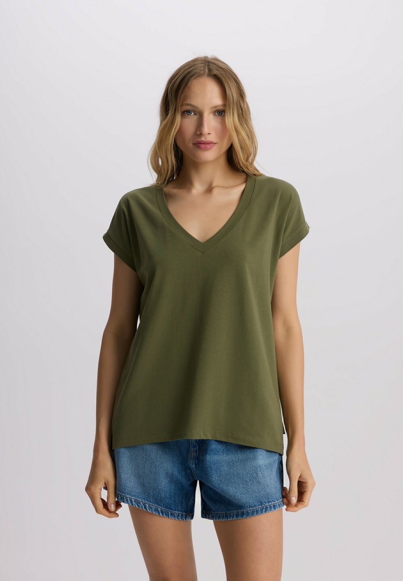 Oliwkowa koszulka typu V-neck z krótkimi, podwiniętymi rękawami, wykonana z gładkiego, lekkiego materiału; zestawiona z dżinsowymi szortami.
