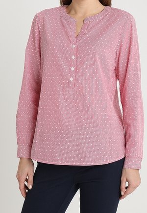 Kobieta ubrana w różową bluzkę z długim rękawem z pionowymi białymi paskami i czterema guzikami z przodu, w połączeniu z ciemnymi spodniami.