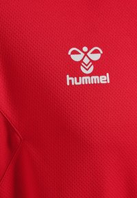 Rød sportstrøje med tekstureret mesh-stof, med et hvidt "hummel" logo og et særpræget geometrisk design ved kraven.
