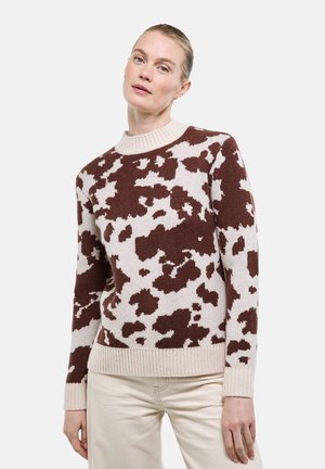 Femme portant un pull en maille marron et beige à motif avec des poignets et un ourlet côtelés, associée à un pantalon beige clair, debout devant un fond uni.