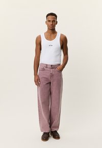 Λευκό αμάνικο μπλουζάκι με μαύρο λογότυπο, συνδυασμένο με ροζ τζιν παντελόνι με φαρδιά πατζάκια και ψηλή μέση, και καφέ δετά παπούτσια. Ουδέτερο φόντο.