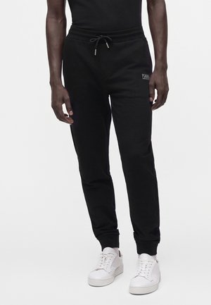 Persona que lleva pantalones jogger negros con cintura ajustable y zapatillas blancas, de pie contra un fondo claro y liso.