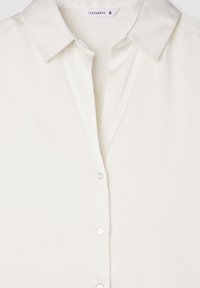 Blusa bianca leggera con colletto, chiusura frontale a bottoni e una consistenza liscia simile alla seta. L'etichetta del marchio è visibile.
