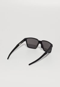 Ochelari de soare negri cu un finisaj mat, ramă unghiulară, lentile întunecate, brațe din cauciuc și un accent metalic pe lateral. Design ușor.