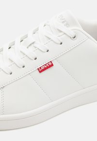Baskets blanches avec une tige synthétique lisse, dotées de coutures ton sur ton, d'une semelle en caoutchouc texturée blanche et d'un logo Levi's rouge sur le côté.