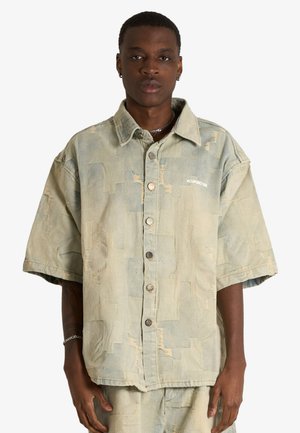 Giovane uomo che indossa una camicia ampia a maniche corte con bottoni e pantaloncini abbinati con un motivo patchwork sottile e testo minimo.