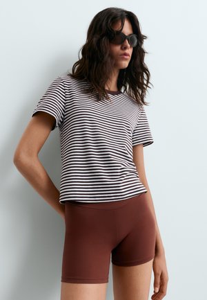 Femme portant des lunettes de soleil foncées, un t-shirt à manches courtes rayé noir et blanc et un short marron ajusté, debout devant un fond clair uni.