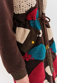 Hnedá fleecová bunda s vzorom nášiviek v červenej, modrej a krémovej farbe, rebrované manžety, predné vrecká s žltými cvokmi a hnedým šnúrkovým uzatváraním.