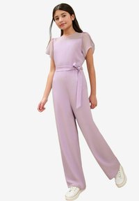 Lavendel-Jumpsuit mit durchsichtigen kurzen Ärmeln, weitem Bein und einem gebundenen Taillengürtel. Kombiniert mit weißen Sneakers mit glatter Textur.