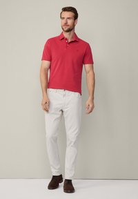 Červené polo tričko s golierom a tromi gombíkmi, spárované s svetlobežovými nohavicami a hnedými topánkami. Jednoduchý dizajn a hladká textúra.