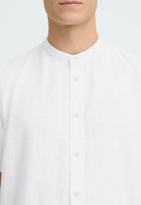 Chemise en lin blanc avec col mao, manches courtes et une rangée de petits boutons beiges sur le devant. Tissu texturé.