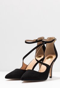 Chaussures à talons hauts en daim noir avec des pointes pointues ; elles présentent des brides croisées à la cheville et un talon aiguille fin. Accent doré sur la semelle intérieure.