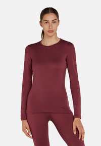 Donna in piedi che indossa un maglione fitted a maniche lunghe color bordeaux e leggings abbinati, rivolta in avanti su uno sfondo bianco uniforme.