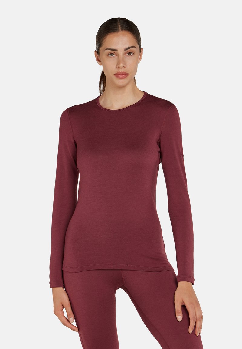 Kvinde står iført en tætsiddende langærmet vinrød top og matchende leggings, vendt fremad mod en ensfarvet hvid baggrund.