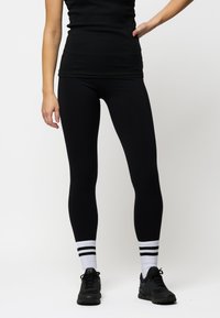 Leggings neri con una superficie liscia, design aderente, abbinati a sneaker nere e calzini a righe bianche. Look semplice e sportivo.