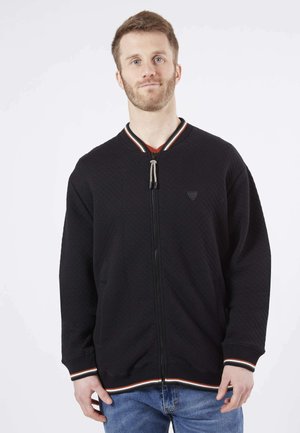 Veste noire matelassée avec une fermeture éclair intégrale, col et poignets côtelés rayés, et un logo sur la poitrine. Comprend deux poches latérales.