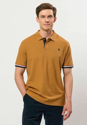 Homme portant un polo jaune moutarde avec un logo bleu marine et des bords de manches rayés, debout avec une main dans la poche sur fond blanc.