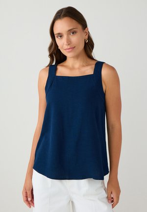 Haut sans manches bleu marine avec encolure carrée et bretelles larges, en tissu texturé, associé à un pantalon blanc.