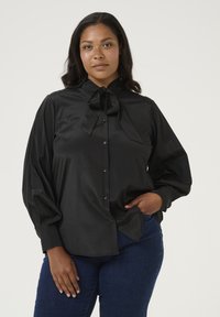 Blusa de satén negra con un alto cuello y un gran lazo en el escote, con mangas largas y holgadas y cierre de botones. Combinada con jeans azules.