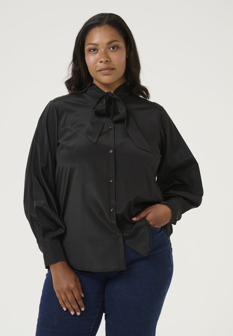 Blusa de satén negra con un alto cuello y un gran lazo en el escote, con mangas largas y holgadas y cierre de botones. Combinada con jeans azules.
