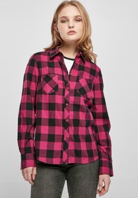 Chemise à carreaux avec de grands carrés roses et noirs, deux poches sur la poitrine, un col et un design à boutons. Le tissu a une texture douce.