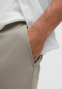 Jasnoszare spodnie wykonane z gładkiej tkaniny, z boczną kieszenią i luźnym krojem. Koszula jest biała z delikatnymi paskami.